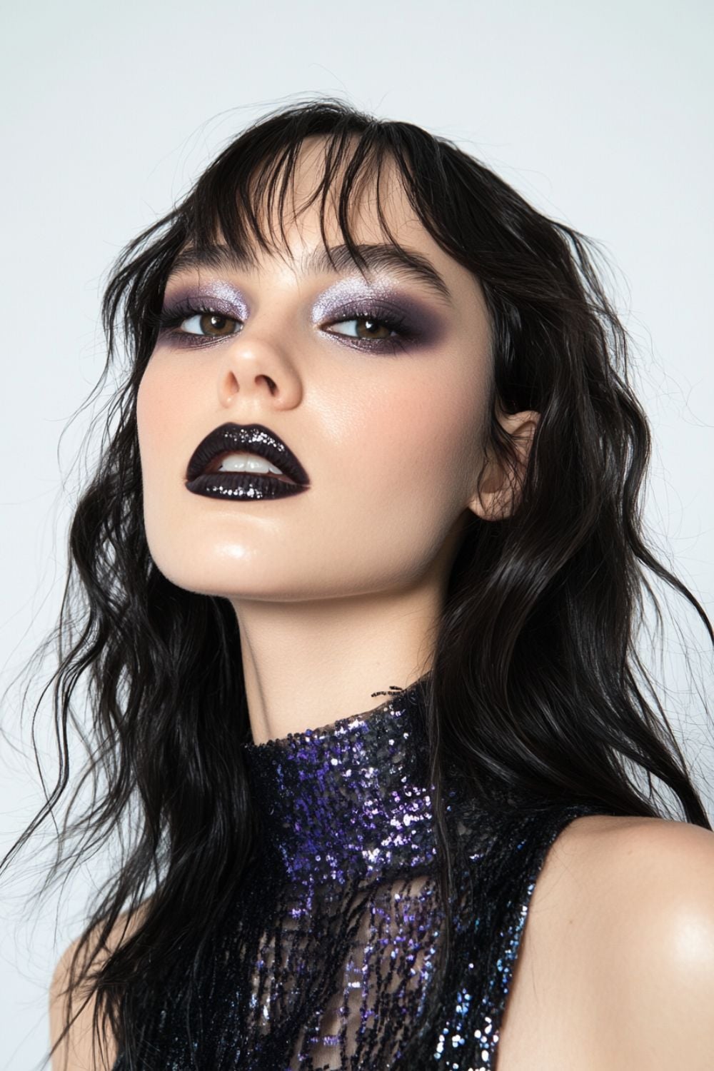 A close-up of a woman with smoky, purple eyeshadow, dramatic winged eyeliner, and glossy black lipstick. She has tousled dark hair and is wearing a sparkling purple sequin top. Her expression is intense and confident as she gazes directly at the camera.